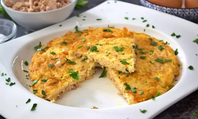 Tortilla de atún dorada y esponjosa, espolvoreada con perejil fresco. Un trozo cortado revela la textura jugosa.