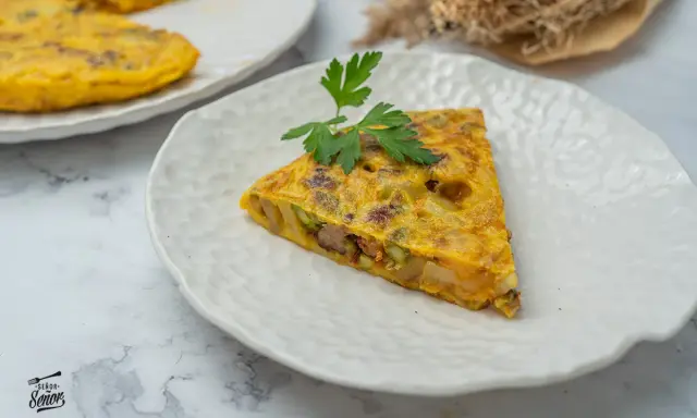Un trozo de tortilla paisana, con patatas, verduras y chorizo, servida en un plato blanco texturizado.