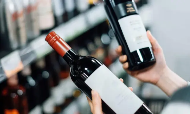 Manos sostienen dos botellas de vinos caros, una con etiqueta blanca y otra con etiqueta negra, en un pasillo de supermercado.