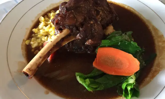 Delicioso lechazo guisado con hueso, acompañado de maíz tierno, zanahoria y verduras verdes en un plato blanco.