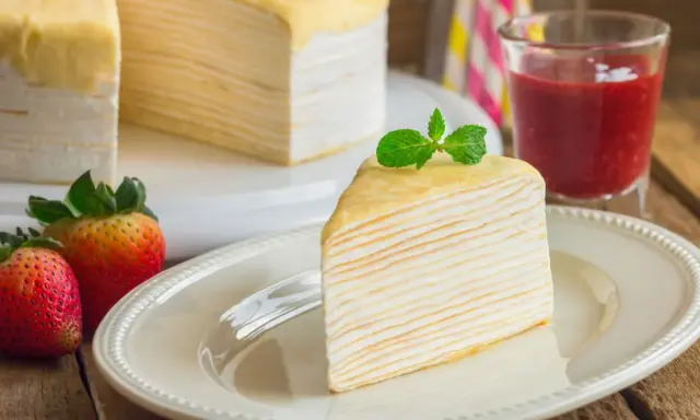 Una porción de tarta de crepes, decorada con menta, junto a fresas y un vaso de batido de fresa.