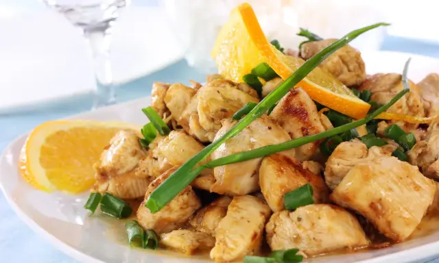 Sabroso pollo a la naranja, trozos jugosos con rodajas de naranja fresca y cebolleta picada, servido en un plato blanco.
