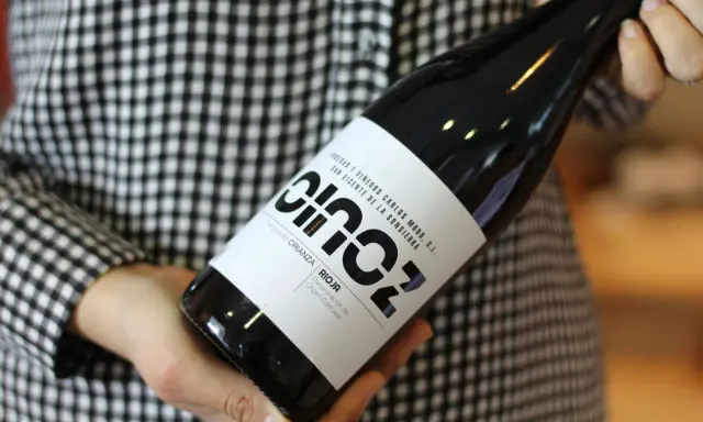 Manos sostienen una botella de vino blanco fermentado en barrica, con etiqueta negra y blanca, sobre un fondo de cuadros.
