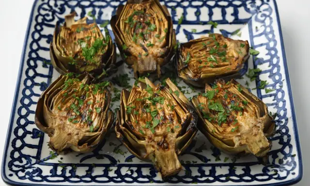 Seis mitades de alcachofas a la plancha, doradas y espolvoreadas con perejil fresco, servidas en un plato decorado.