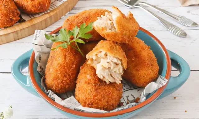 Croquetas de atún doradas y crujientes, servidas en un cuenco azul. Una está partida, mostrando su cremoso interior.