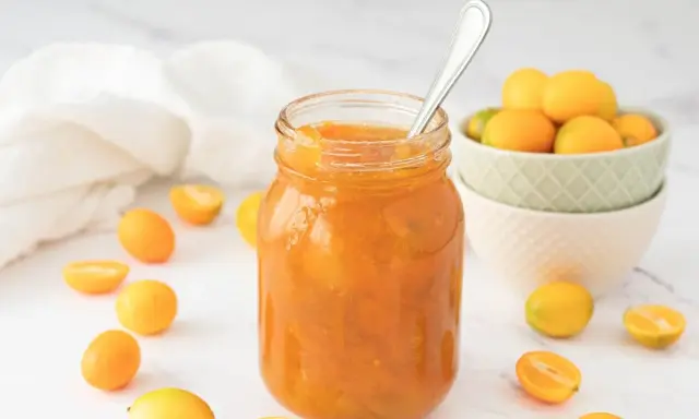 Tarro de mermelada de kumquat casera, rodeado de kumquats frescos y cortados. Una cuchara plateada reposa en el tarro.