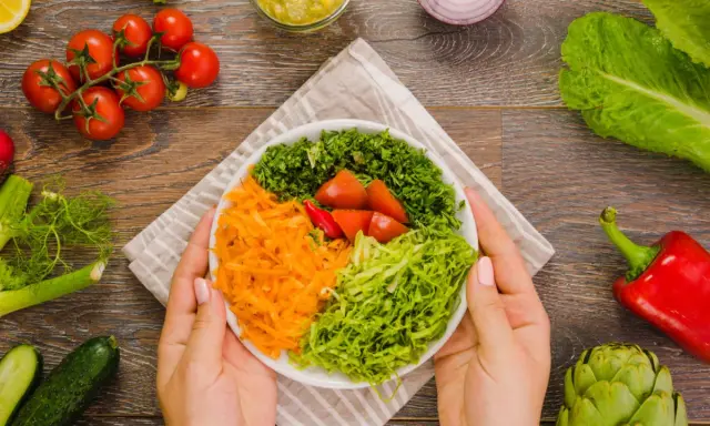 Manos sostienen un plato con una colorida ensalada mediterránea: zanahoria rallada, lechuga, perejil picado y tomates.