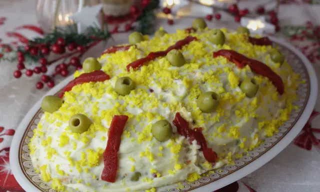 Ensaladilla rusa, la receta de la abuela, cubierta de mayonesa, huevo rallado, aceitunas y pimientos. ¡Un clásico festivo!