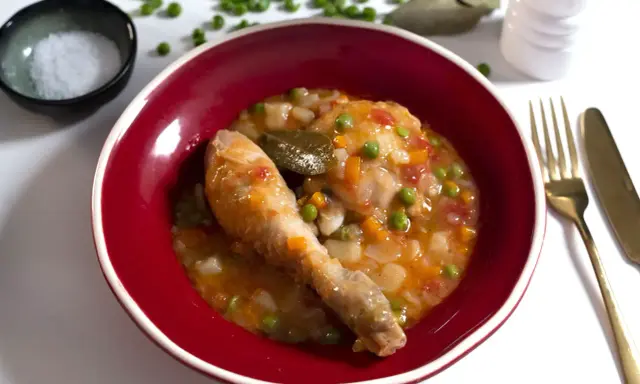 Un delicioso pollo a la jardinera en un cuenco rojo, con guisantes, zanahoria y tomate. Acompañado de sal, laurel y cubiertos dorados.