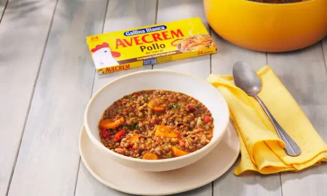 Un tazón de lentejas con zanahoria y pimiento rojo, listo para servir. La caja de Avecrem Pollo de corral, clave en esta lentejas receta de la abuela, descansa al lado.