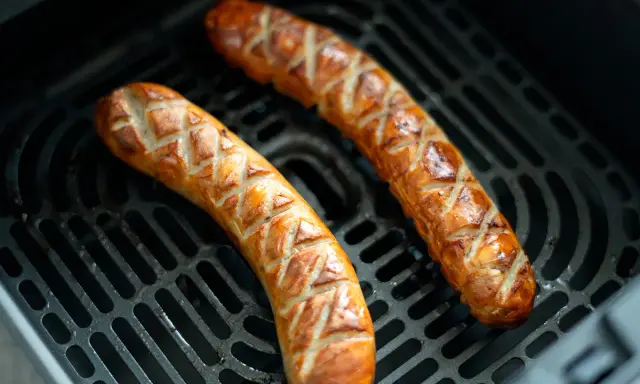 Dos salchichas doradas con cortes en forma de rombos, cocinándose en la rejilla de una freidora de aire.
