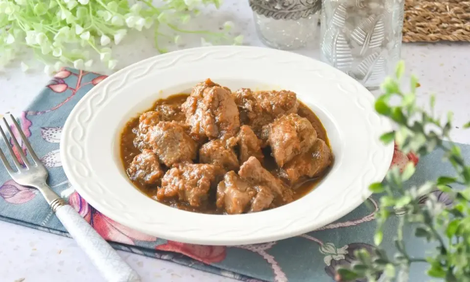 Plato de tierna carne de cerdo en salsa, servido en un cuenco blanco con un tenedor y decoración floral.