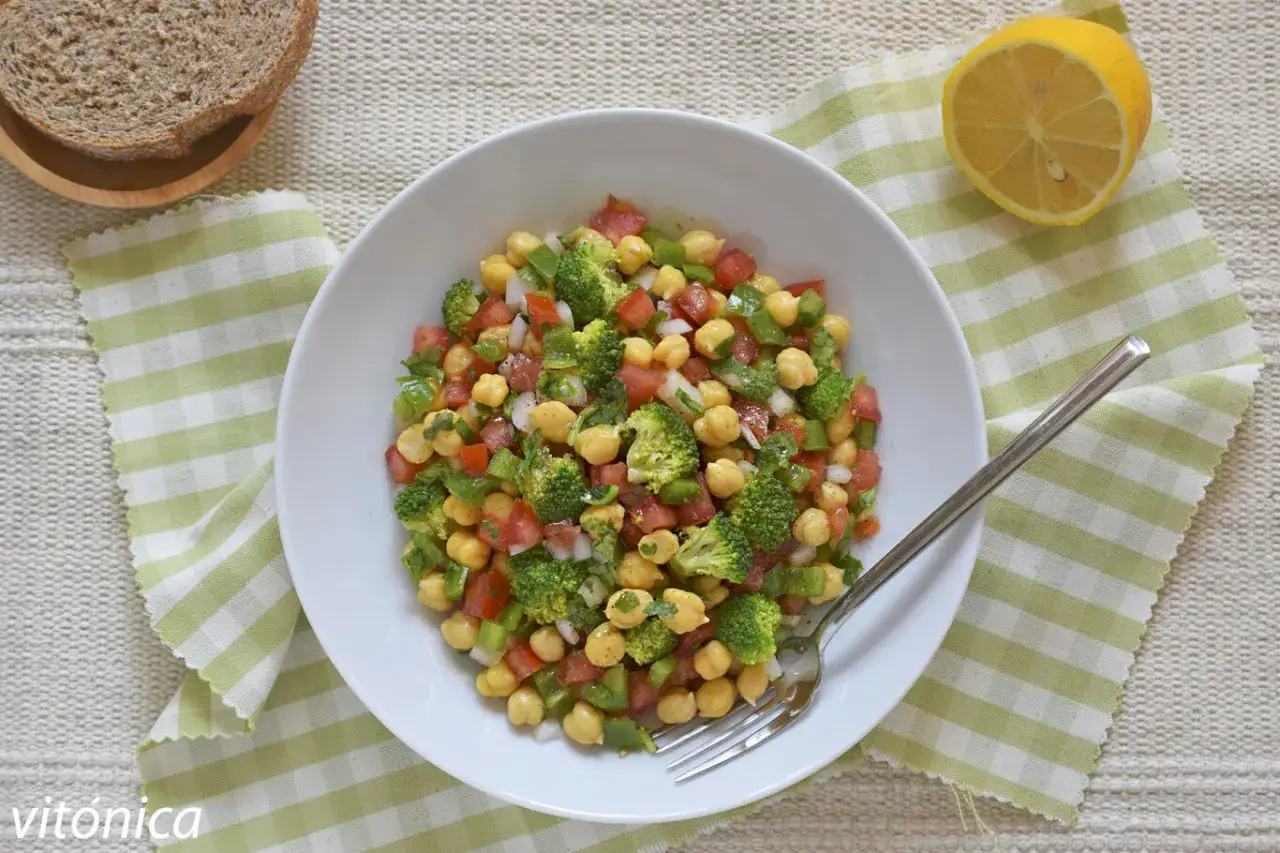 Ensalada garbanzos vibrante con br&oacute;coli, tomate y pimiento verde. Servida en un bol blanco con pan y lim&oacute;n al lado.