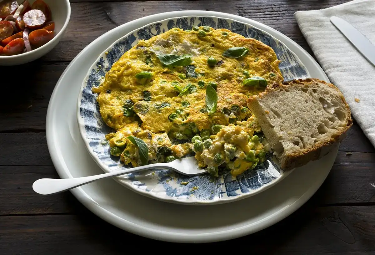 Tortilla de guisantes, esponjosa y dorada, servida con pan r&uacute;stico y una ensalada de tomates.