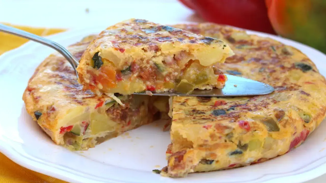 Una porci&oacute;n de tortilla paisana, jugosa y llena de verduras como pimientos y calabac&iacute;n, se levanta con una esp&aacute;tula.