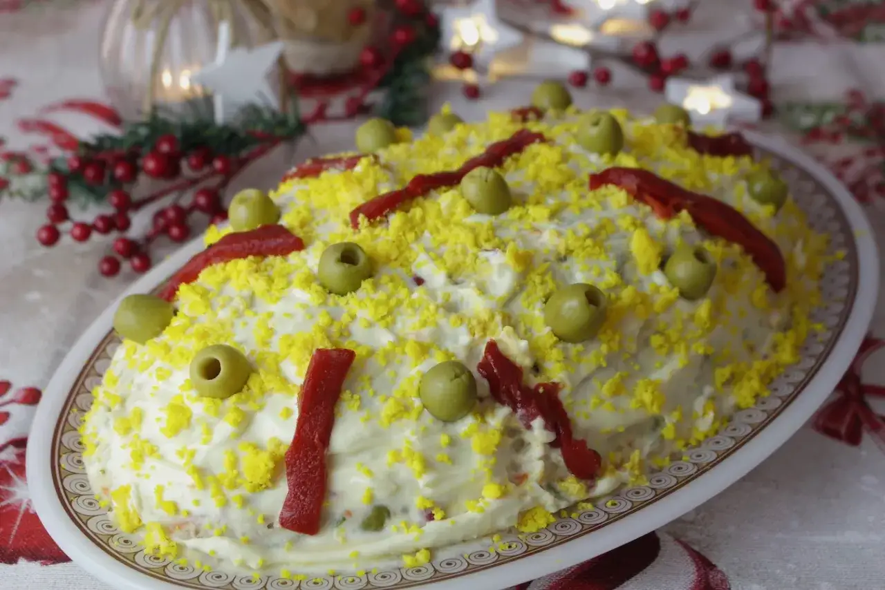 Ensaladilla rusa, la receta de la abuela, cubierta de mayonesa, huevo rallado, aceitunas y pimientos.
