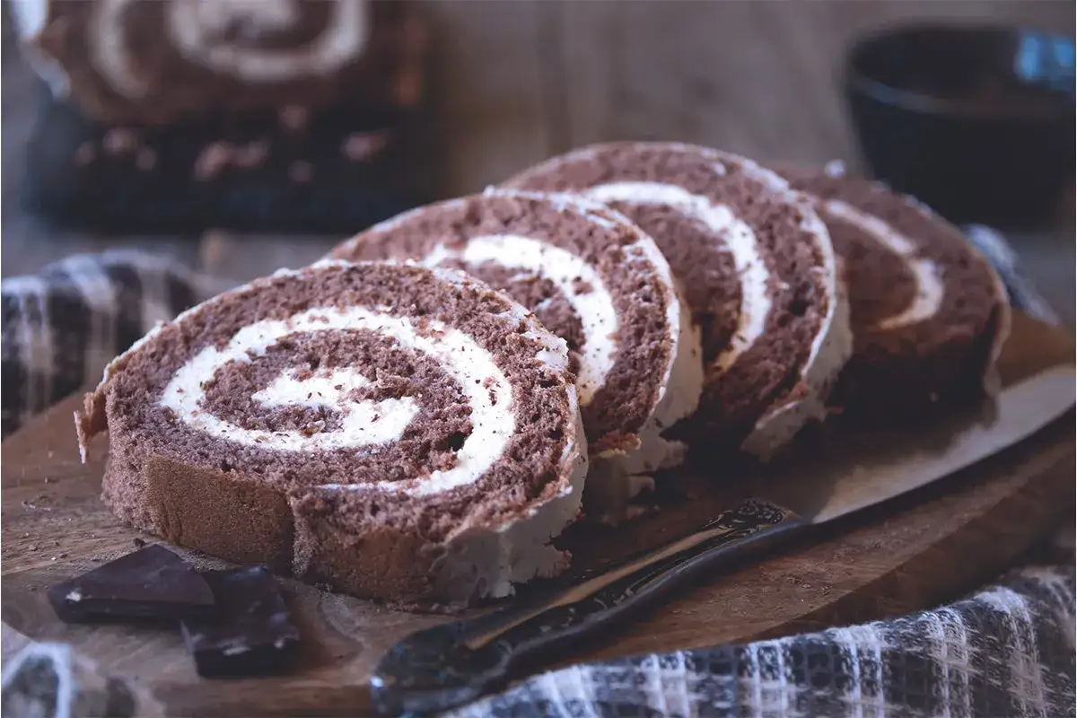 Un delicioso rollo de chocolate con crema blanca, cortado en rodajas sobre una tabla de madera.