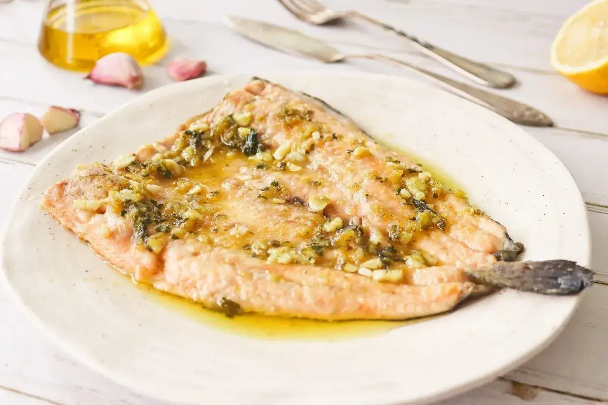 Trucha al ajillo servida en plato blanco, con aceite de oliva, ajos y lim&oacute;n de fondo.