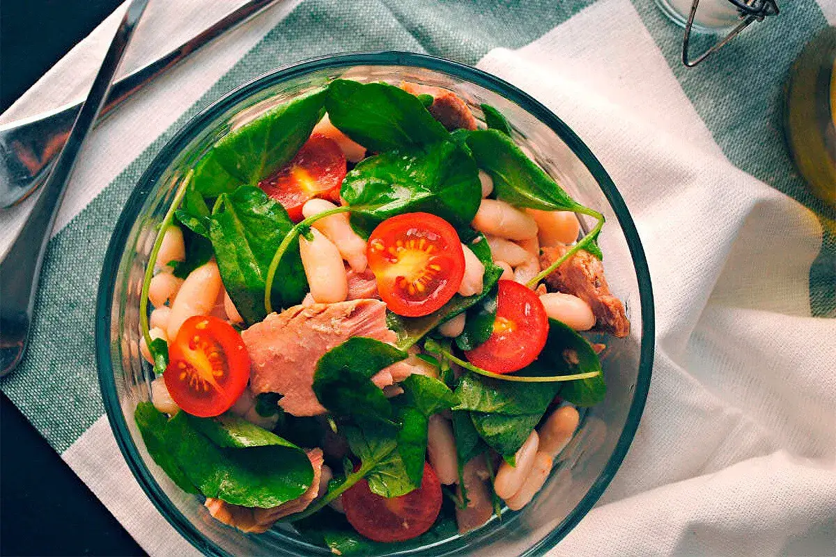 Ensalada de alubias con at&uacute;n, espinacas y tomates cherry, servida en un bol de cristal.