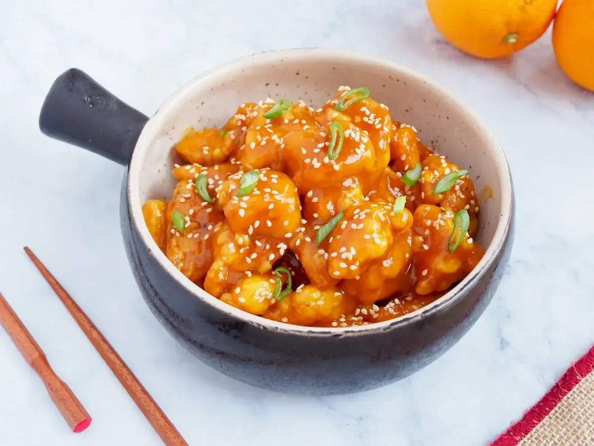 Un delicioso pollo a la naranja, cubierto con semillas de s&eacute;samo y cebolleta, servido en un cuenco con palillos.