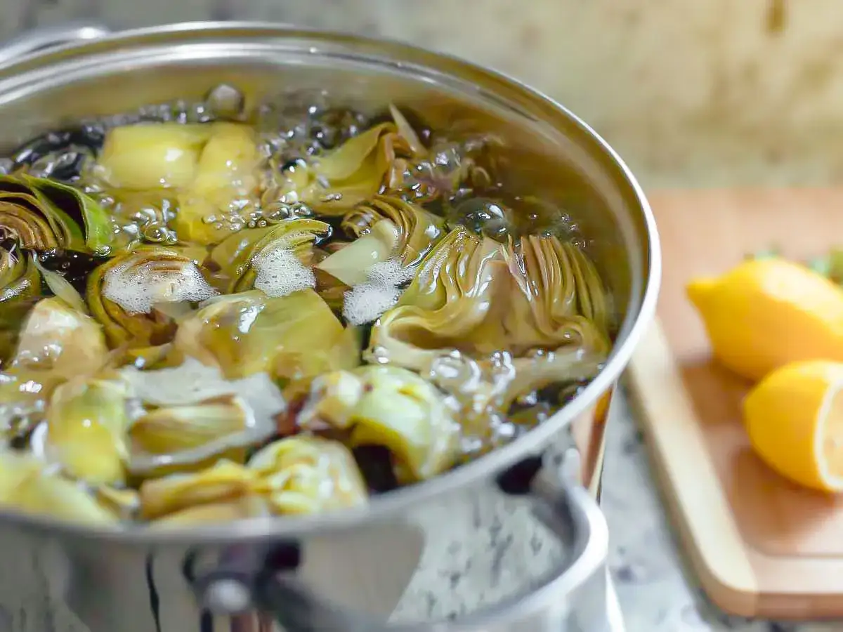 Alcachofas cortadas hirviendo en una olla, un paso clave para aprender como cocer alcachofas. Limones en una tabla de cortar al lado.