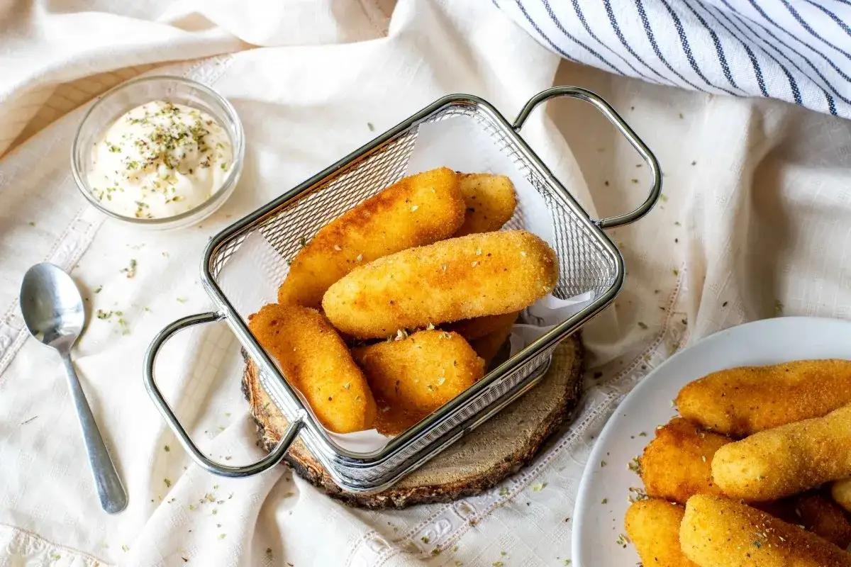 Croquetas de patata doradas y crujientes en una cesta met&aacute;lica, acompa&ntilde;adas de una salsa cremosa.
