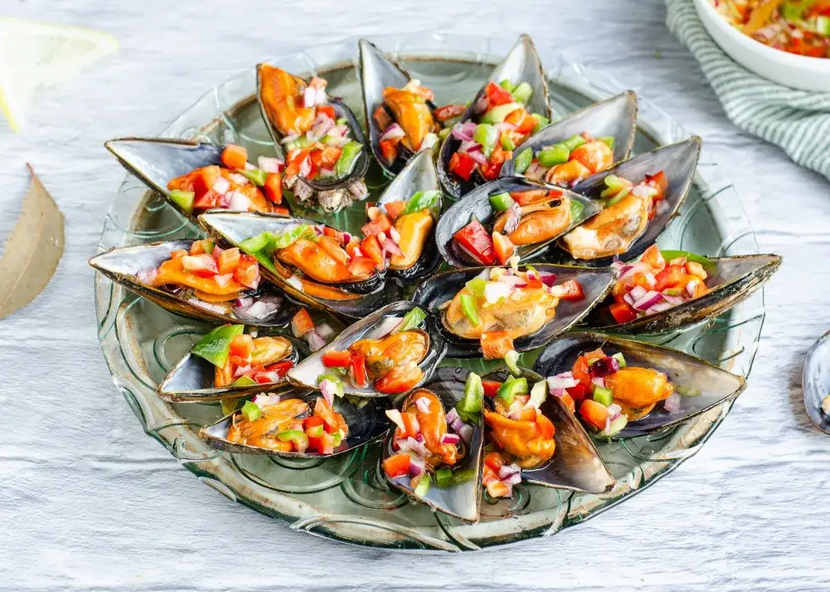 Mejillones a la vinagreta servidos en una bandeja de cristal, adornados con una mezcla de pimientos y cebolla picados.