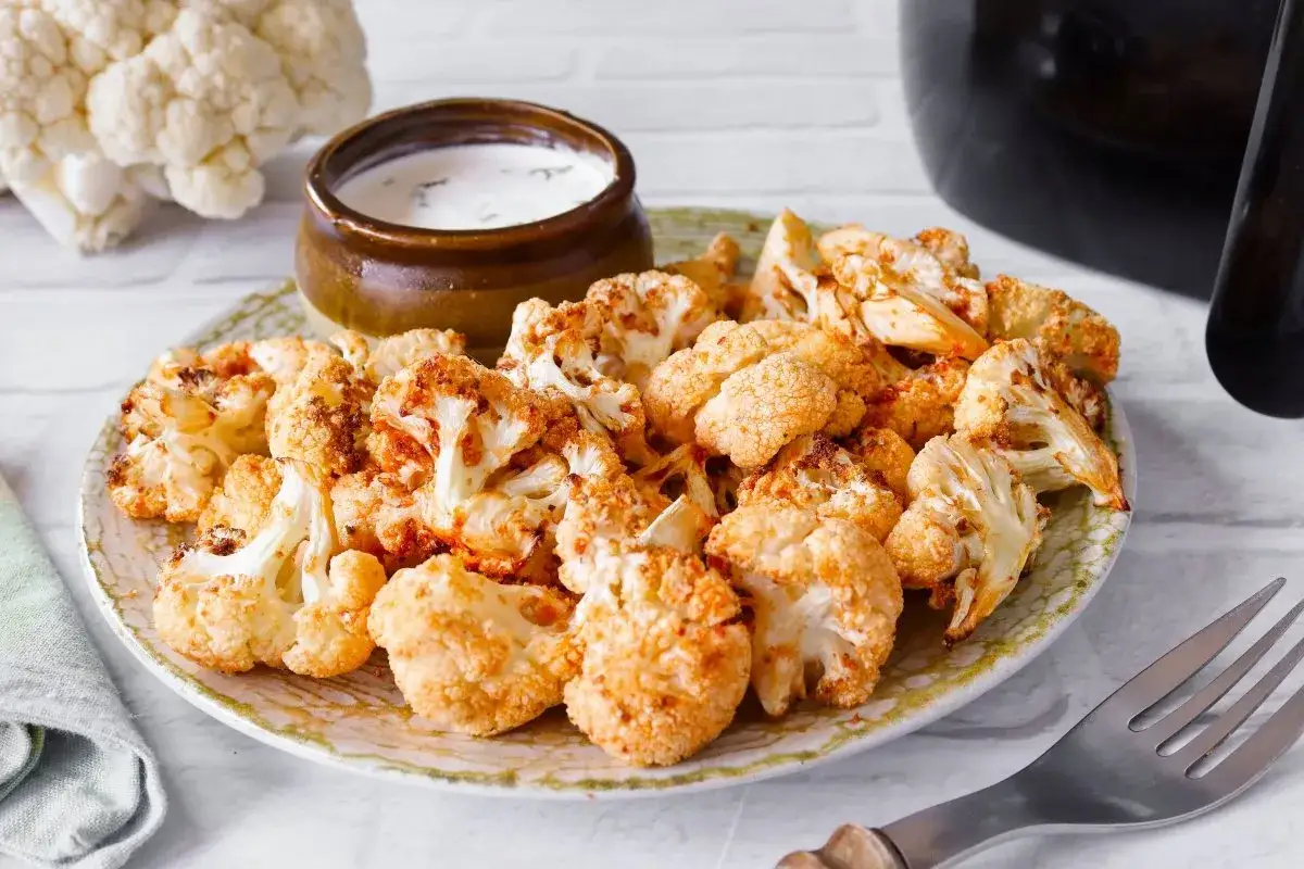 Coliflor airfryer crujiente y dorada, servida en un plato con un taz&oacute;n de salsa cremosa.