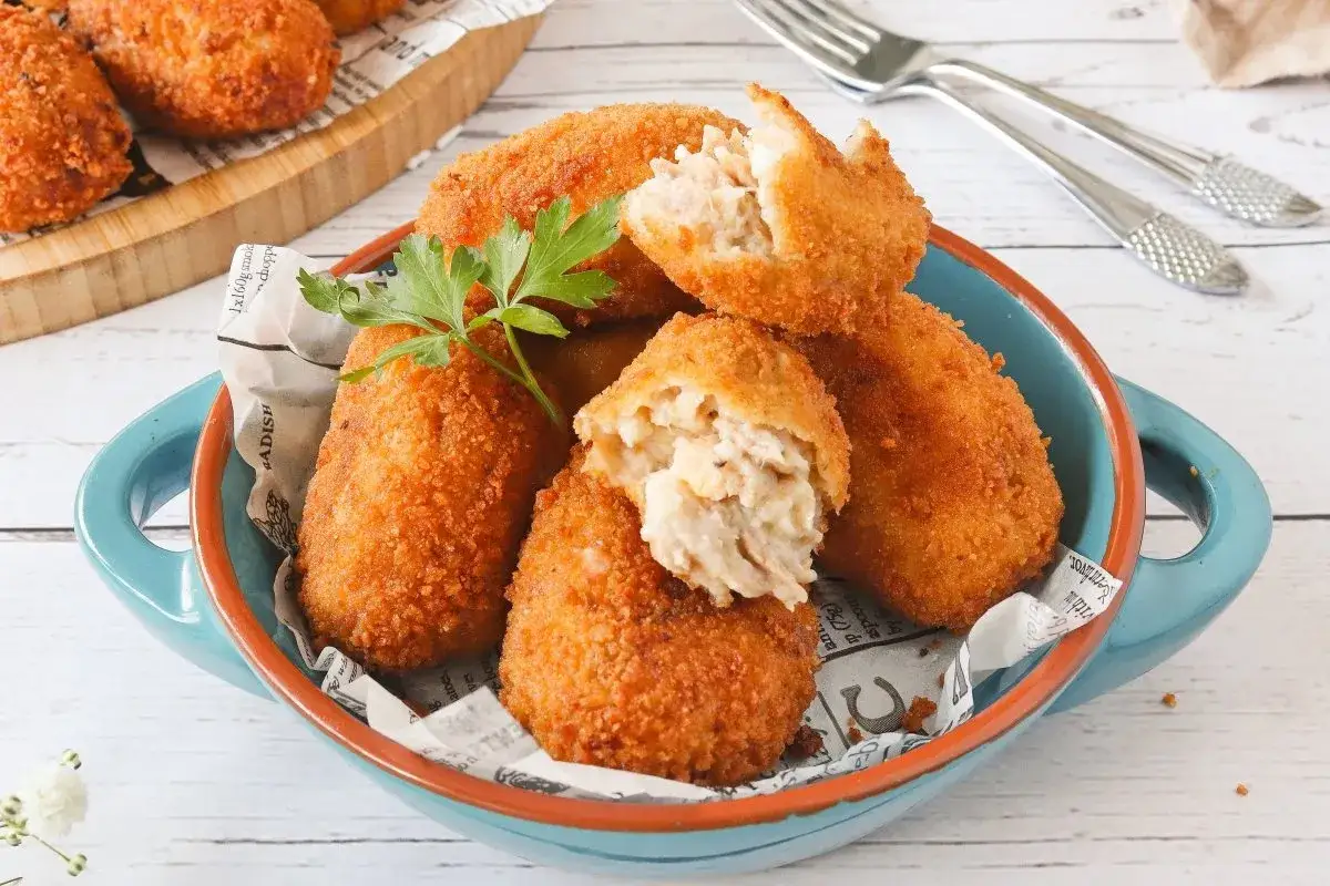 Croquetas de at&uacute;n doradas y crujientes, servidas en un cuenco azul. Una est&aacute; partida, mostrando su cremoso interior.