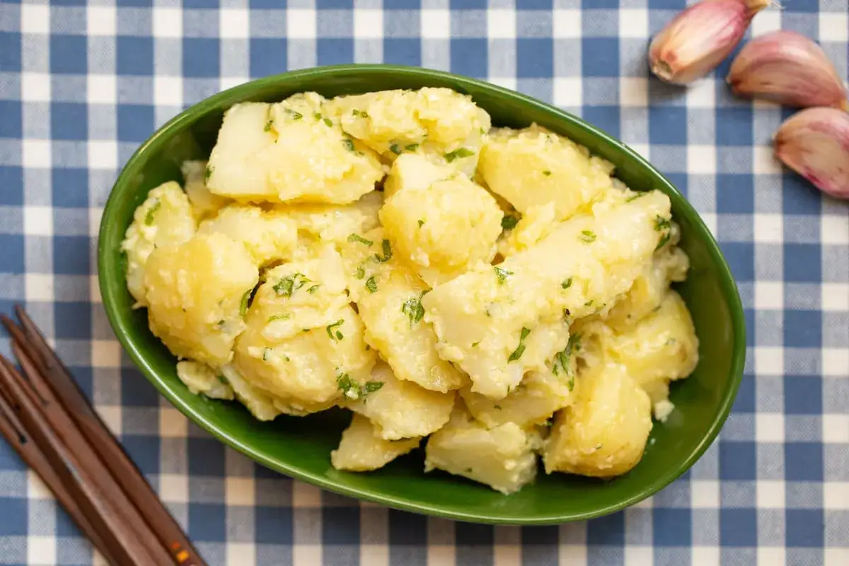 Un cuenco verde lleno de patatas alioli cremosas, espolvoreadas con perejil fresco. A su lado, unos dientes de ajo y unos palillos de madera.