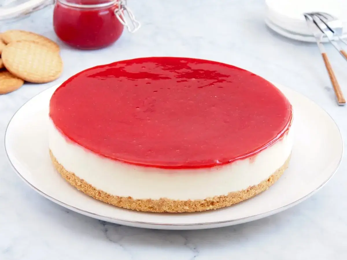 Tarta de queso Philadelphia con cobertura roja brillante, base de galleta y un tarro de mermelada de fresa al fondo.