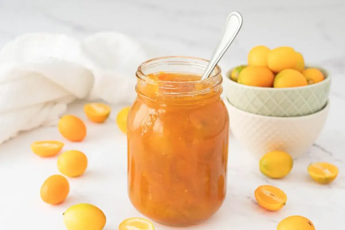 Tarro de mermelada de kumquat casera, rodeado de kumquats frescos y cortados. Una cuchara plateada reposa en el tarro.