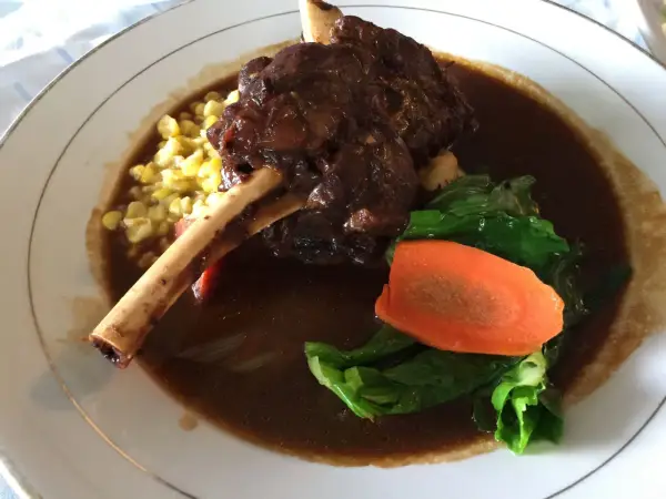 Delicioso lechazo guisado con hueso, acompañado de maíz tierno, zanahoria y verduras verdes en un plato blanco.