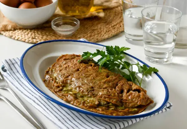 Tortilla de alcachofas dorada y jugosa, servida en plato blanco con borde azul. Decorada con perejil fresco.