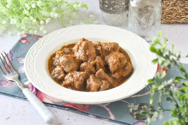 Plato de tierna carne de cerdo en salsa, servido en un cuenco blanco con un tenedor y decoración floral.