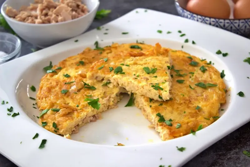 Tortilla de atún dorada y esponjosa, espolvoreada con perejil fresco. Un trozo cortado revela la textura jugosa.