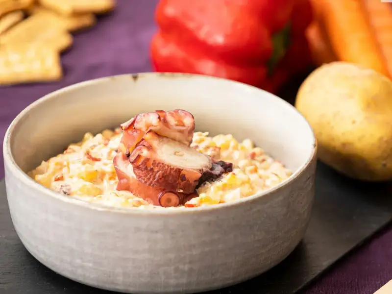 Deliciosa ensaladilla de pulpo cremosa, servida en cuenco, con patata, pimiento y zanahoria de fondo.